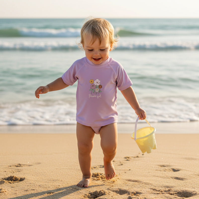 Baby läuft am Strand und trägt ein mauvefarbenes Bade-T-Shirt mit Blumenmotiv – sommerliche Baby-Bademode am Meer