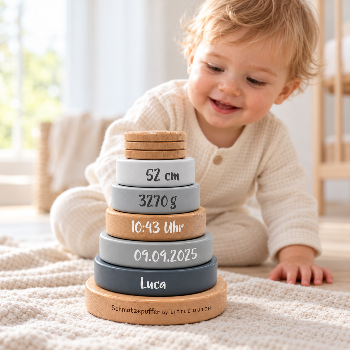 Grauer Holz-Stapelturm mit Name Luca und Geburtsdaten auf einer cremefarbenen Decke; ein Kind sitzt dahinter im hellen Kinderzimmer.