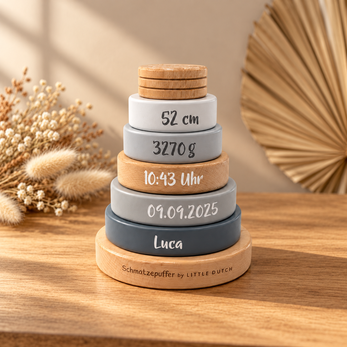 Gepersonaliseerde houten stapeltoren met de naam Luca en geboortegegevens, in grijs en naturel hout, warm verlicht naast droogbloemen.